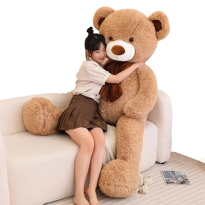 Young woman hugging an oversized brown teddy bear plush while sitting on a white sofa, showing the large, soft, cuddly design of the toy.